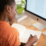 Best Remote Jobs for Indian Students 2026: Earn ₹10,000-50,000/Month While Studying A woman in a polka-dot shirt writes in a notebook at her desk with a computer screen visible.